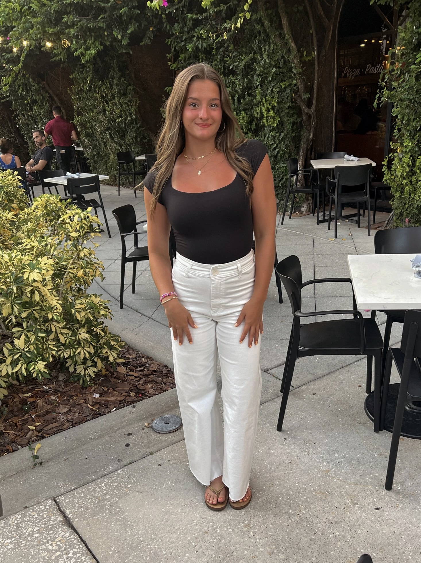 Color photograph of Ava Jackowsky, a white female with light brown hair and brown eyes, standing smiling at the camera. She is wearing a black cap sleeve top and white jeans with flip flops. The background is the outdoor seating area of a restaraunt in Sarasota, Florida.